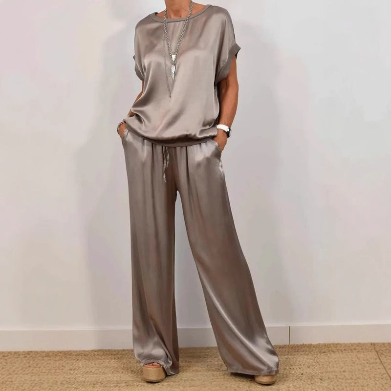 Donna in completo satinato beige, composto da maglia a maniche corte e pantaloni larghi. Indossa sandali con zeppa e collana lunga. Moda elegante.