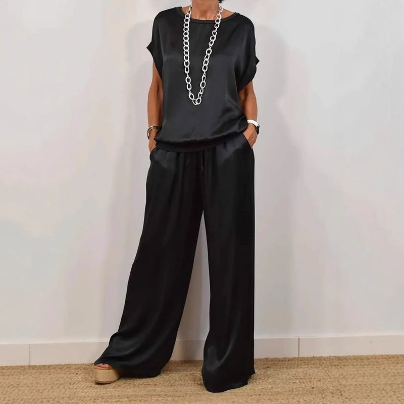 Donna in elegante completo nero di seta, composto da maglia e pantaloni larghi, indossa collana lunga e sandali con zeppa. Moda femminile chic.