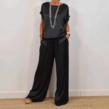 Donna in elegante completo nero di seta, composto da maglia e pantaloni larghi, indossa collana lunga e sandali con zeppa. Moda femminile chic.