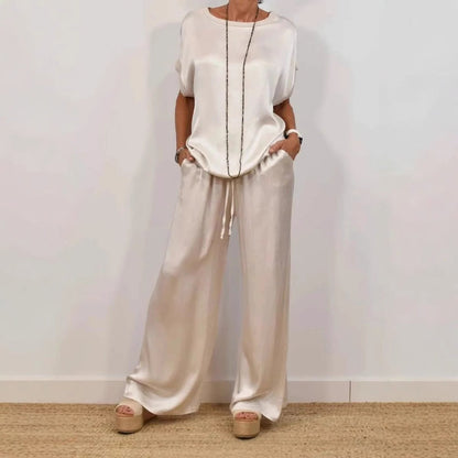 Donna in abbigliamento elegante, indossa un completo bianco di seta con pantaloni larghi e top morbido, accessoriato con collana lunga.
