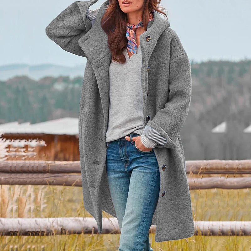 Donna con cappotto grigio invernale, jeans e sciarpa colorata, in un paesaggio rurale. Moda autunnale, abbigliamento casual, stile outdoor.