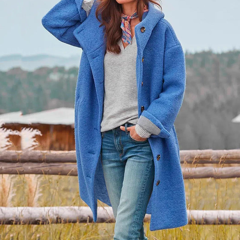 Donna con cappotto blu, maglione grigio e jeans, posa all'aperto davanti a una staccionata di legno. Sfondo naturale autunnale.