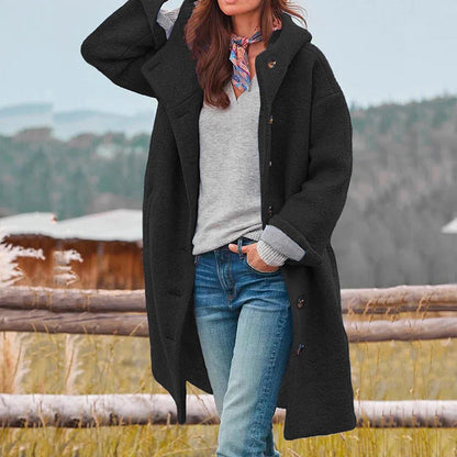 Donna con cappotto nero lungo, jeans e sciarpa colorata, in posa all'aperto davanti a una staccionata di legno, sfondo naturale autunnale.