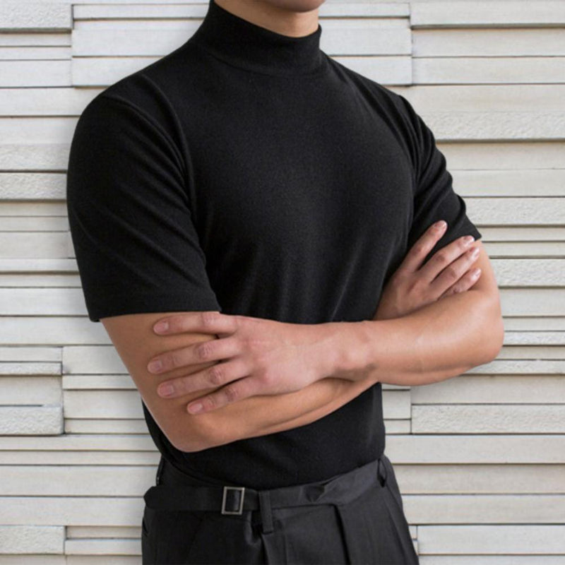 Uomo in maglia nera a collo alto, braccia incrociate, sfondo di pannelli bianchi. Moda elegante, abbigliamento maschile, stile minimalista.