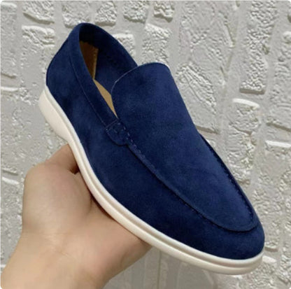Mocassino blu scamosciato da uomo, suola bianca, tenuto in mano, sfondo muro bianco. Scarpe eleganti, moda maschile, calzature casual.