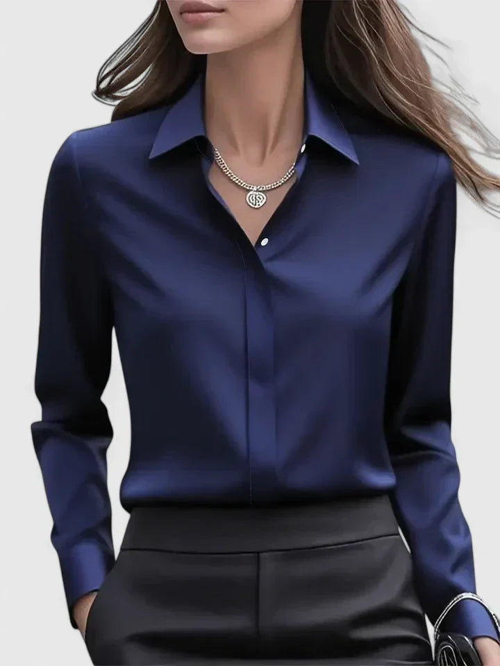 Donna in camicia di seta blu scuro, elegante e lucida, con collana d'argento, sfondo grigio chiaro. Moda femminile, abbigliamento formale.