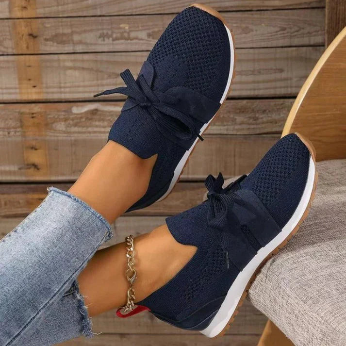 Scarpe da ginnastica blu scuro con fiocco, suola bianca e dettagli in maglia, indossate con jeans chiari. Design casual e moderno per donna.