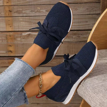 Scarpe da ginnastica blu scuro con fiocco, suola bianca e dettagli in maglia, indossate con jeans chiari. Design casual e moderno per donna.