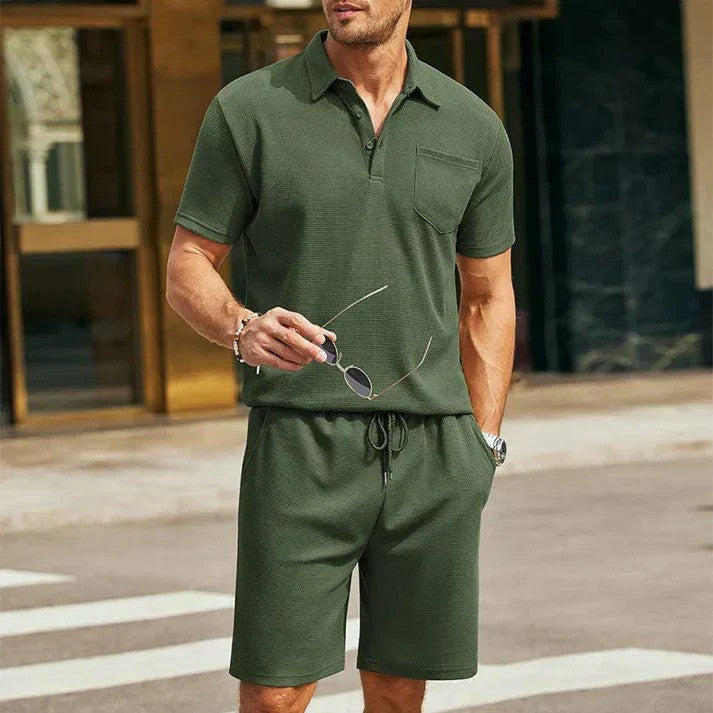 Uomo in completo estivo verde oliva, camicia polo e pantaloncini, tiene occhiali da sole. Moda casual maschile, abbigliamento estivo elegante.