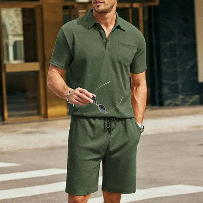 Uomo in completo estivo verde oliva, camicia polo e pantaloncini, tiene occhiali da sole. Moda casual maschile, abbigliamento estivo elegante.