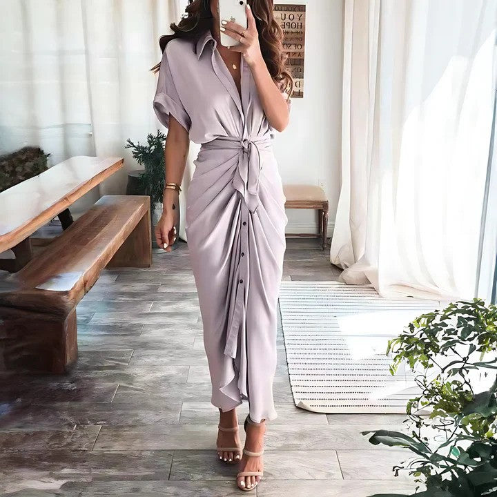 Donna in abito lungo elegante color lavanda con nodo frontale, sandali con tacco, in un salotto moderno con tavolo in legno e piante decorative.