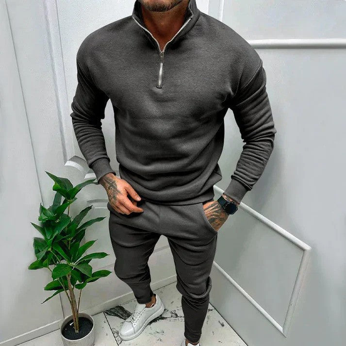 Uomo in tuta sportiva grigia con zip, orologio nero e scarpe bianche, accanto a pianta verde in vaso, in ambiente moderno.