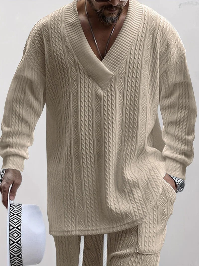 Uomo con maglione beige a trecce, scollo a V profondo, pantaloni coordinati, orologio al polso, cappello bianco con motivo geometrico in mano.