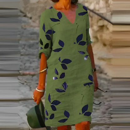 Donna con abito verde a foglie blu, maniche arrotolate, cammina su strada acciottolata. Moda estiva, stile casual, accessori argento.