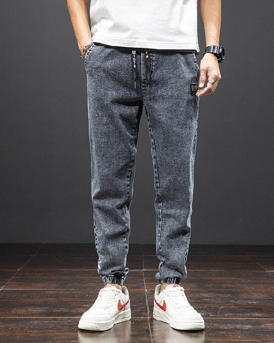 Jeans jogger uomo grigio scuro, stile casual, con elastico alla caviglia, abbinati a scarpe da ginnastica bianche con dettagli rossi.