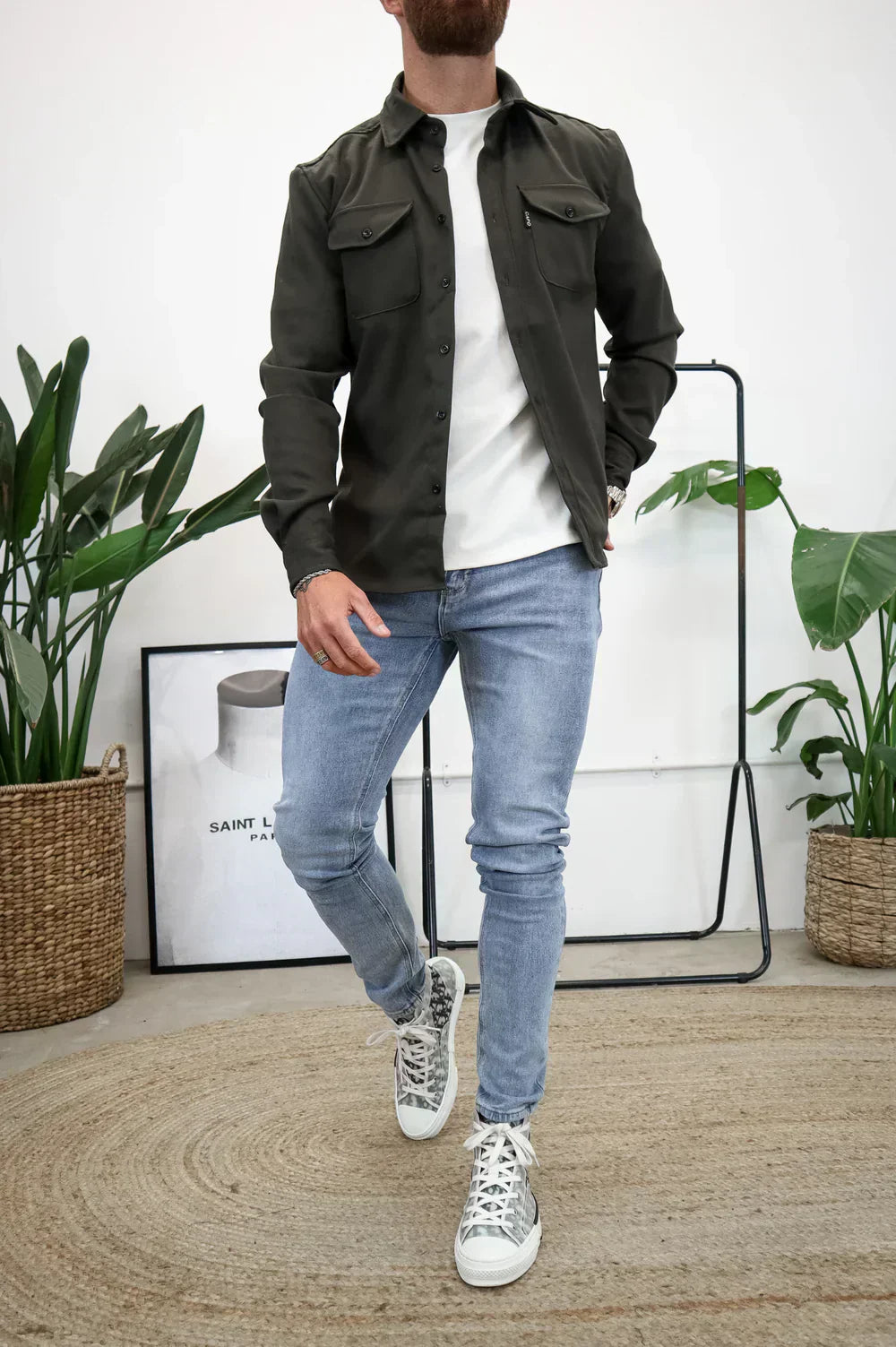 Uomo in camicia verde militare e jeans chiari, posa casual in interni moderni con piante e poster. Moda maschile, stile urbano.