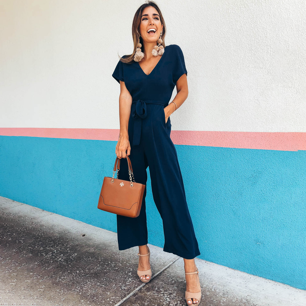 Donna sorridente in tuta blu elegante, con borsa marrone e sandali beige, posa davanti a muro colorato. Moda femminile, abbigliamento estivo chic.