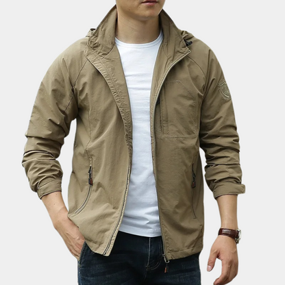 Giacca a vento beige da uomo, stile casual, con zip frontale e tasche laterali, indossata su maglietta bianca e jeans. Moda maschile autunnale.