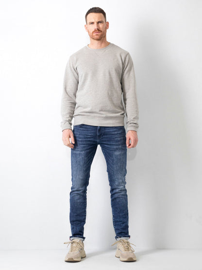 Uomo in piedi con maglione grigio, jeans blu e scarpe beige su sfondo bianco. Moda casual maschile, abbigliamento autunnale, stile moderno.