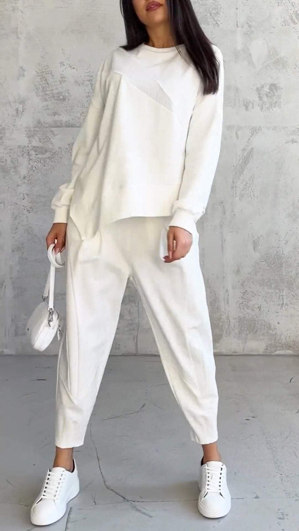 Donna in abbigliamento casual bianco, con maglione e pantaloni coordinati, borsa a tracolla e scarpe da ginnastica, su sfondo grigio.