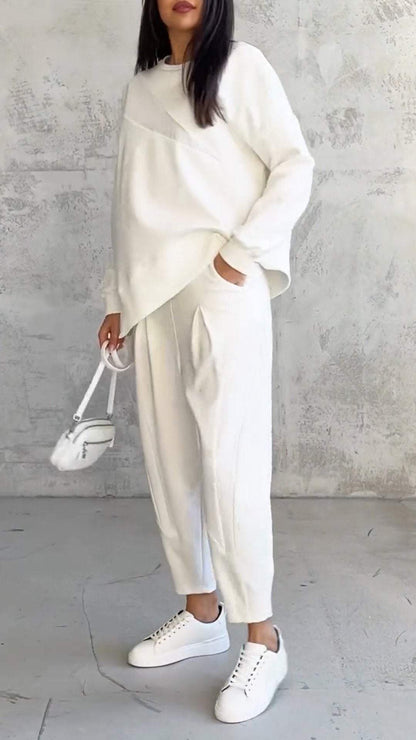 Donna in abbigliamento casual bianco, con maglione oversize e pantaloni, borsa a tracolla e sneakers, su sfondo grigio minimalista.