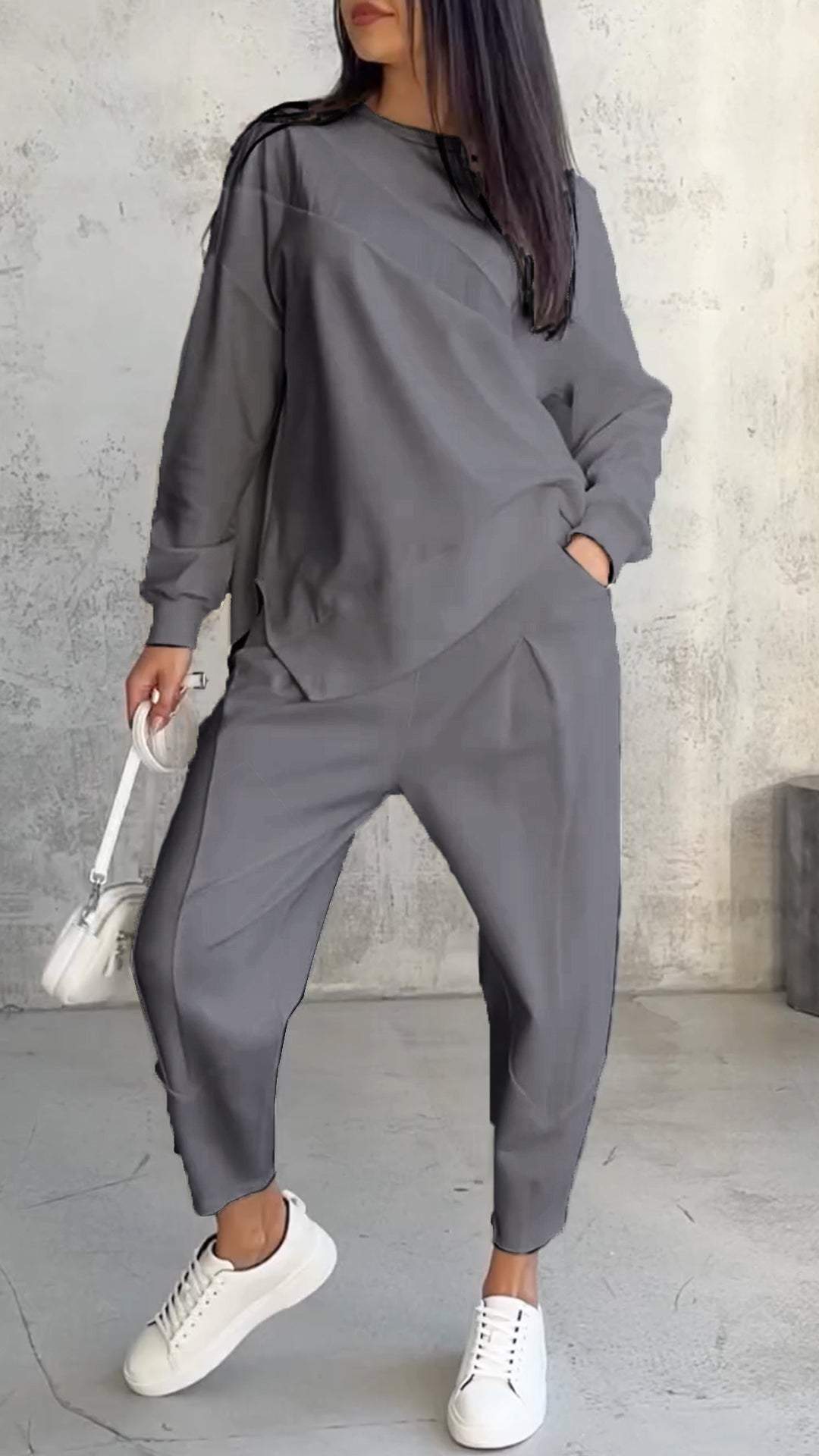 Donna in tuta grigia casual, con borsa bianca e sneakers, posa su sfondo grigio. Moda autunnale, abbigliamento comodo, stile urbano.