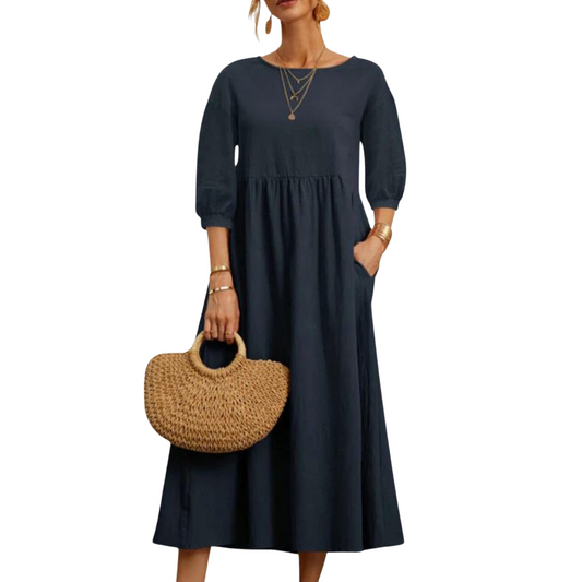 Abito lungo blu scuro con maniche a tre quarti, indossato con borsa in paglia intrecciata e gioielli dorati. Moda donna elegante e casual.