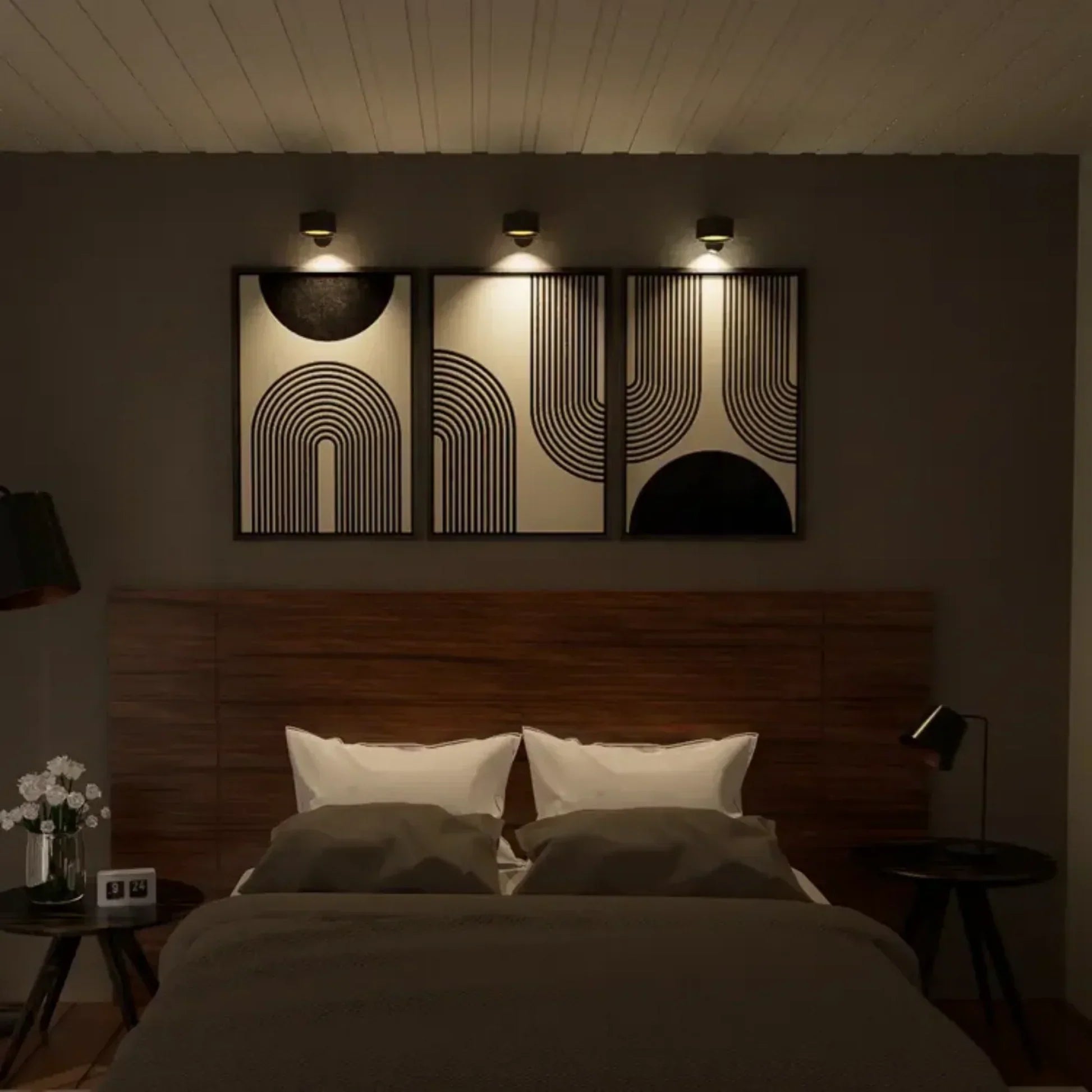 Camera da letto moderna con letto matrimoniale, testiera in legno, cuscini bianchi, coperta grigia, quadri astratti e illuminazione soffusa.