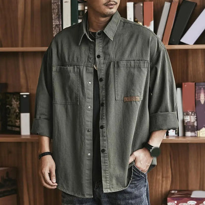 Uomo con camicia verde militare oversize, maniche arrotolate, davanti a una libreria. Moda casual maschile, stile urbano, tendenze abbigliamento.