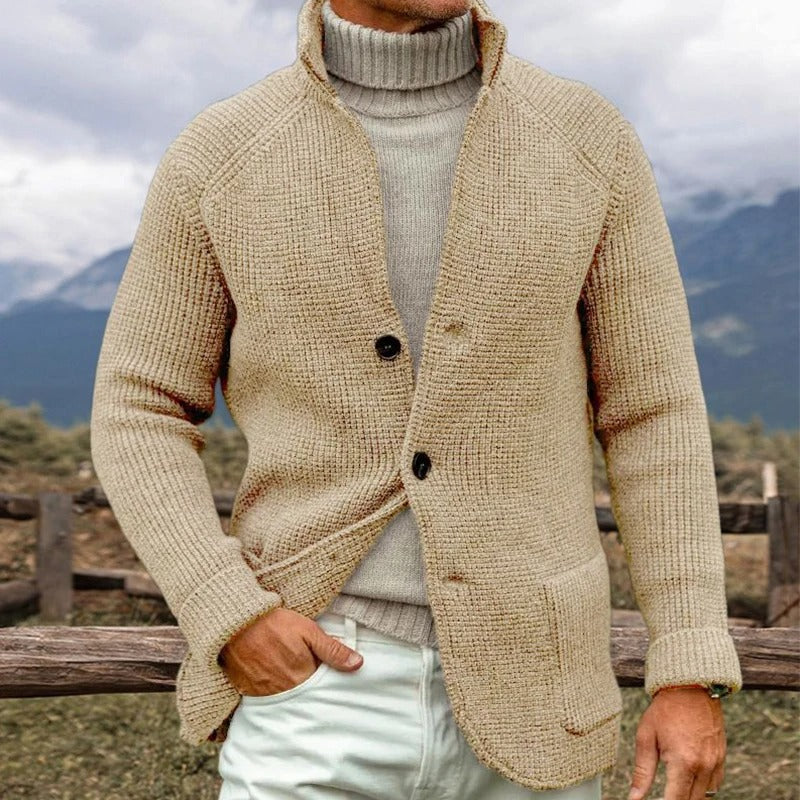Uomo con maglione beige a collo alto e cardigan in lana, sfondo montano. Moda autunnale, abbigliamento caldo, stile casual elegante.