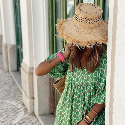Donna con abito verde a motivi geometrici e cappello di paglia, appoggiata a un muro bianco. Moda estiva, stile boho chic, accessori casual.