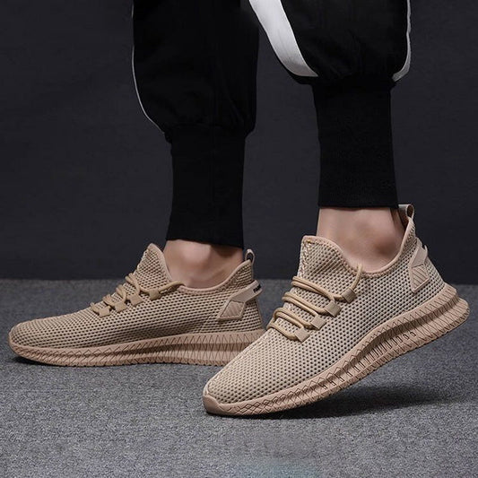 Scarpe da ginnastica beige su sfondo grigio, indossate con pantaloni sportivi neri. Design moderno, suola robusta, perfette per attività sportive e casual.