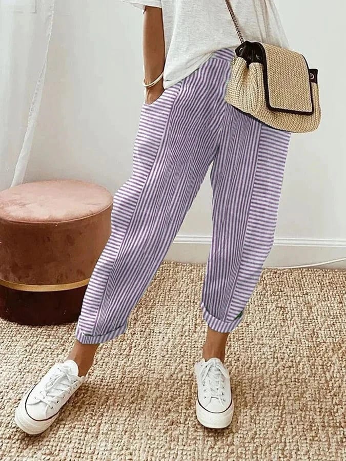 Donna con pantaloni a righe viola e bianche, borsa a tracolla in paglia, maglietta bianca e sneakers bianche su tappeto intrecciato.