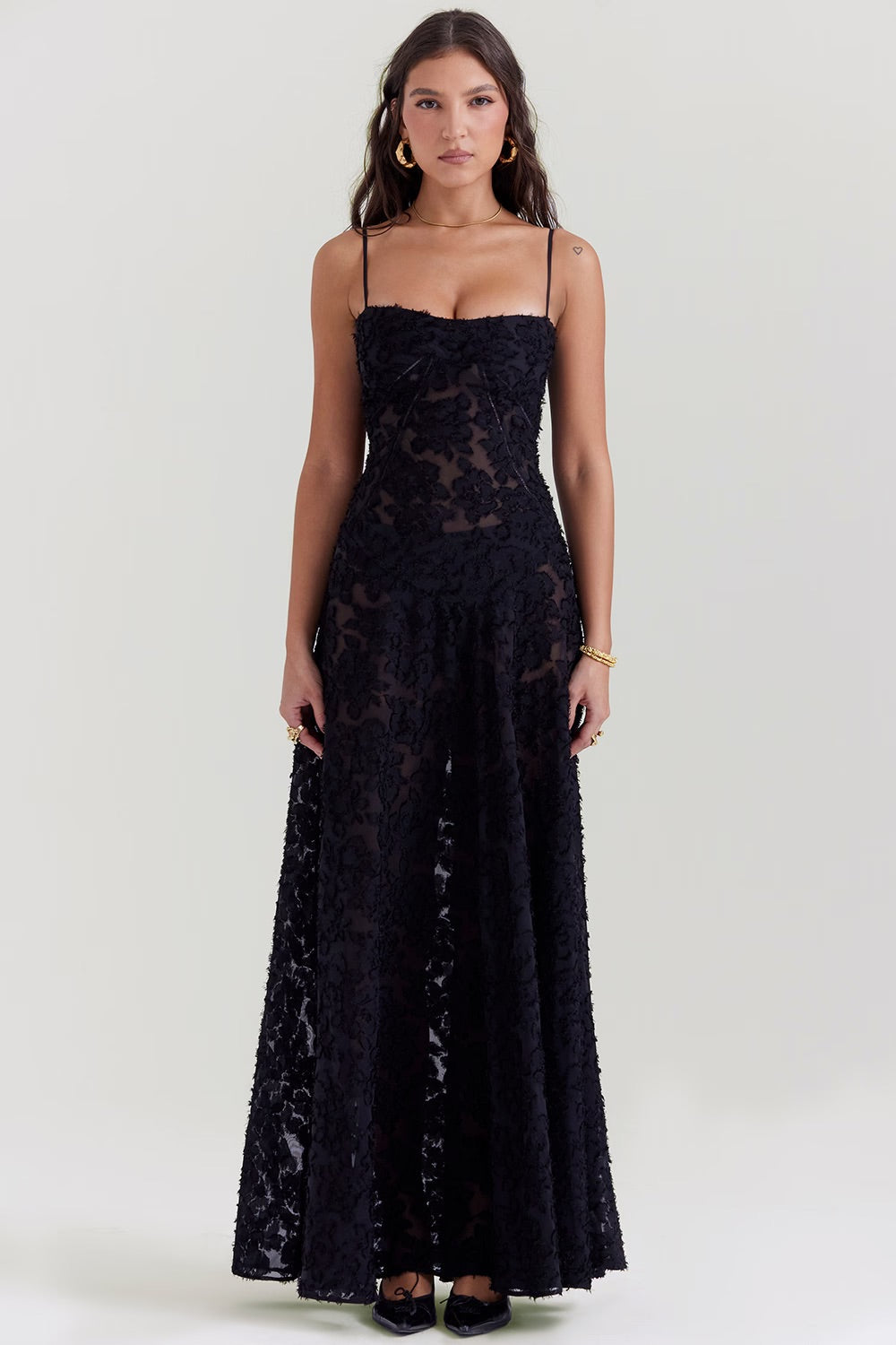 Donna in abito lungo nero trasparente con motivi floreali, spalline sottili, orecchini dorati e bracciale. Moda elegante, abbigliamento serale.