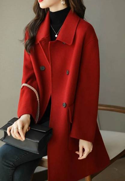 Donna con cappotto rosso elegante, seduta su sedia, indossa maglione nero e tiene borsa nera. Moda autunnale, stile chic, abbigliamento femminile.