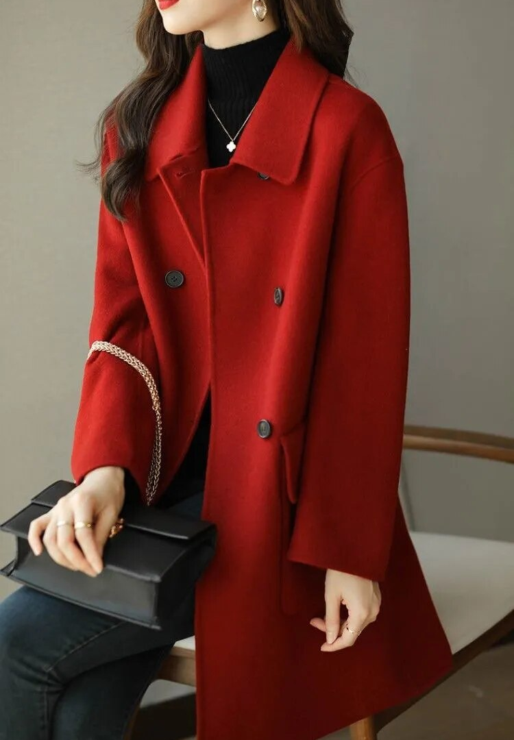 Donna con cappotto rosso elegante, seduta su sedia, indossa maglione nero e orecchini, tiene borsa nera. Moda autunnale, stile chic.