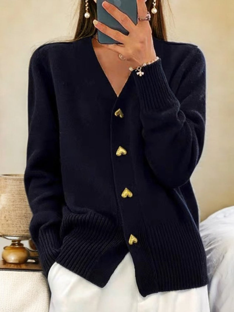 Donna con cardigan nero con bottoni a forma di cuore, in piedi accanto a un letto e una lampada. Stile casual elegante, moda autunnale.