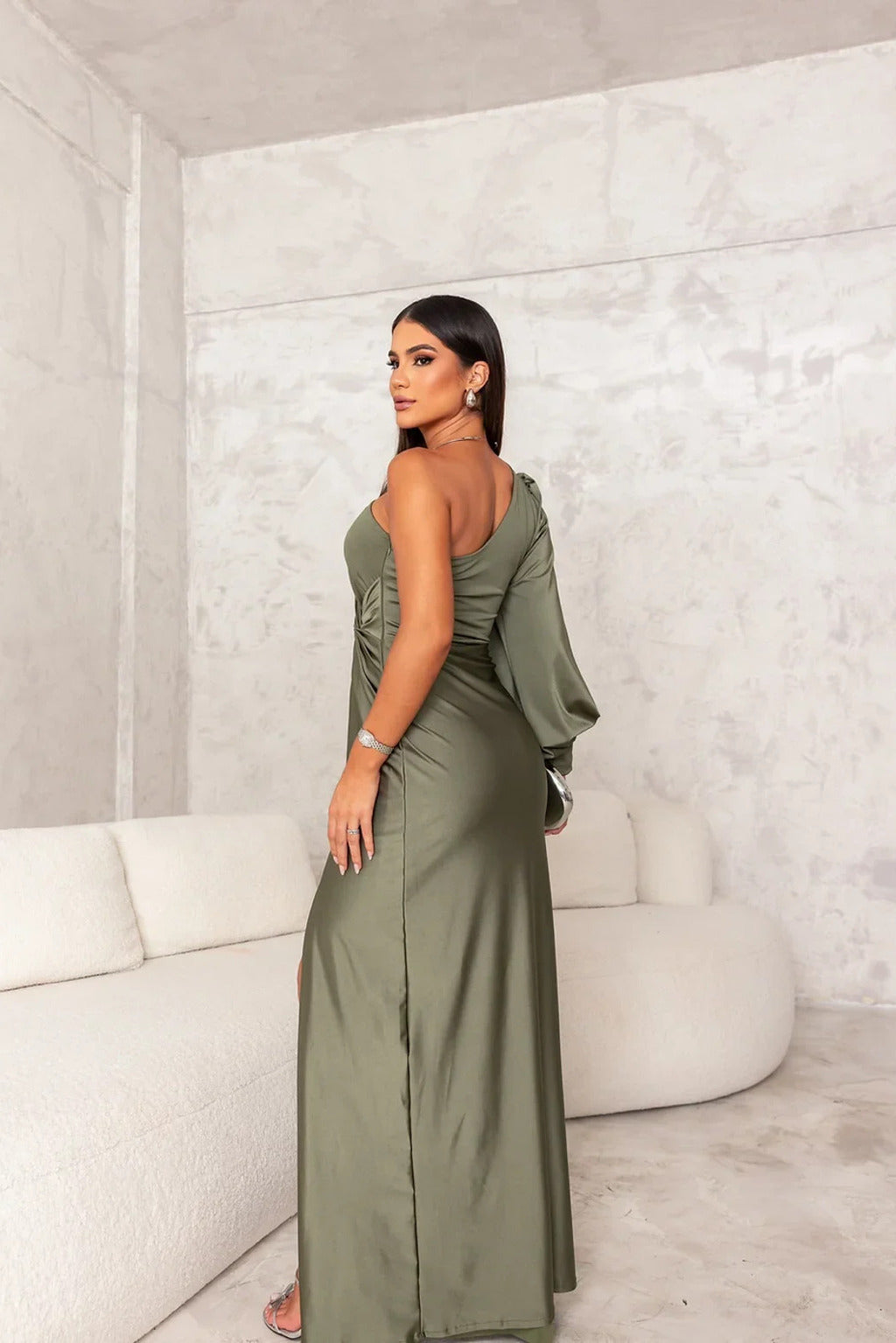 Donna in abito lungo verde oliva elegante, con spacco laterale, in posa in un salotto moderno con divano bianco. Moda femminile, abbigliamento elegante.
