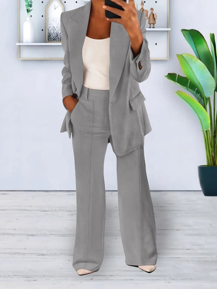 Donna in elegante completo grigio, giacca e pantaloni, in posa moderna. Sfondo con pianta verde e mensola decorativa. Moda femminile contemporanea.