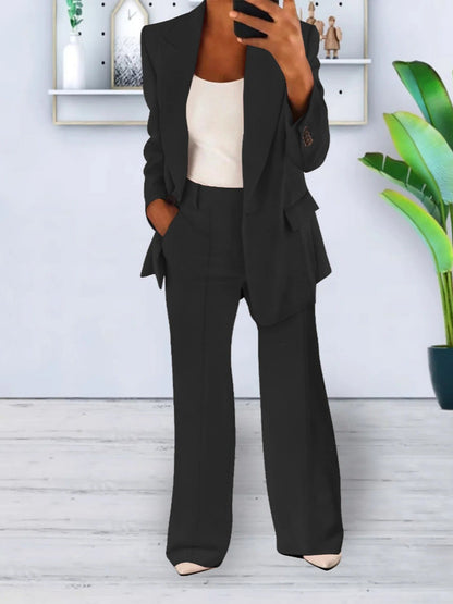 Donna in completo nero elegante, giacca e pantaloni, con top bianco. Sfondo moderno con pianta verde e scaffali decorativi. Moda business casual.