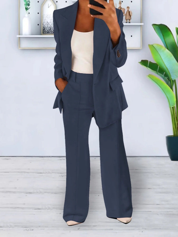 Donna in elegante completo blu scuro, giacca e pantaloni, con top bianco. Sfondo moderno con pianta verde e decorazioni minimaliste.