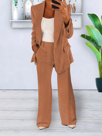 Donna in elegante completo pantalone marrone, top bianco, in piedi su pavimento in legno, con piante e scaffali moderni sullo sfondo.