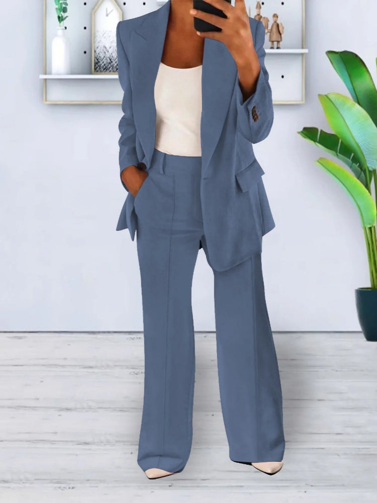 Donna in completo blu elegante, giacca e pantaloni, con top bianco. Interno moderno con pianta verde e mensole sullo sfondo. Moda femminile formale.